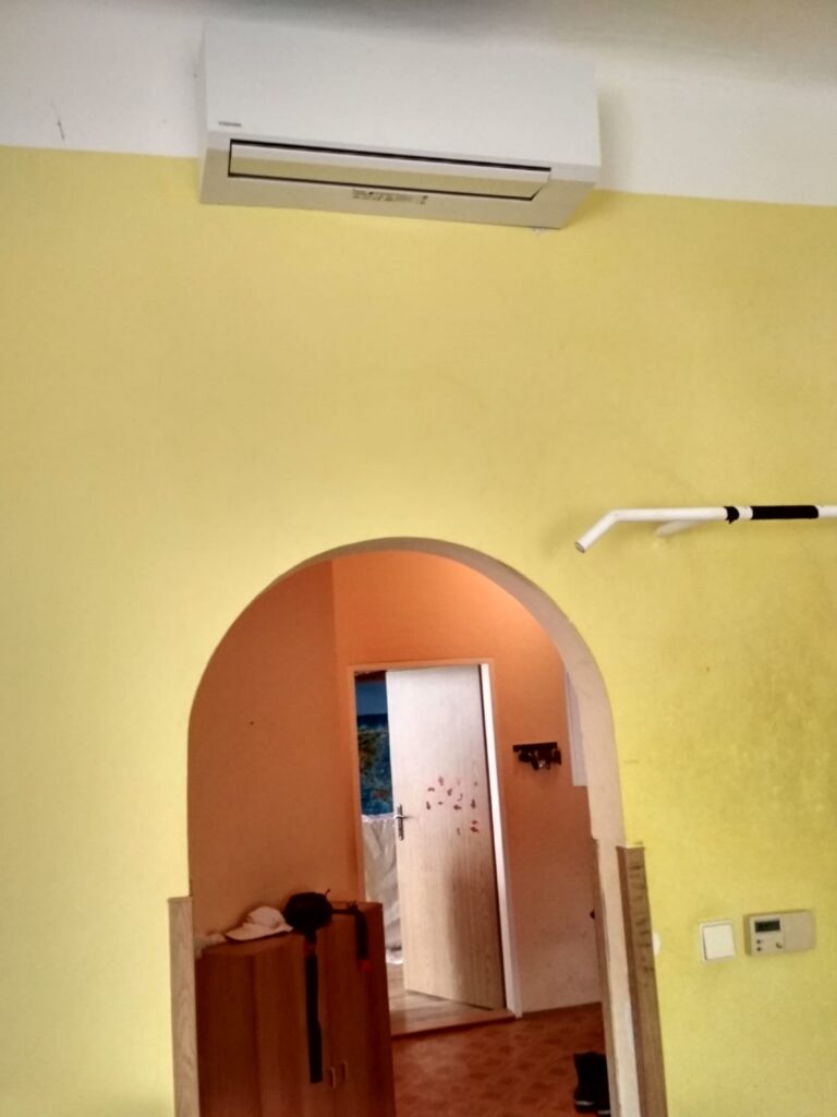 Nástěnná klimatizační jednotka nad obloukovým průchodem v místnosti se žlutou stěnou